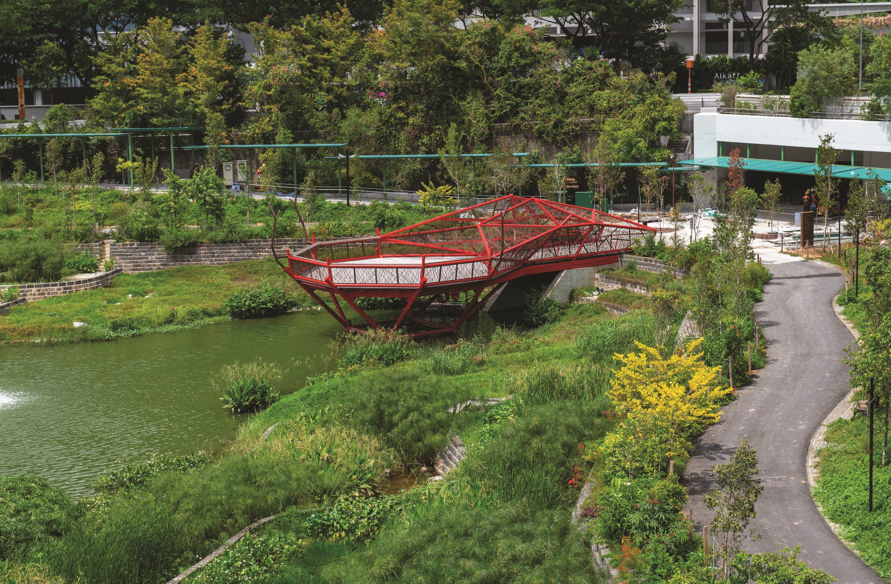 An elevated view of Alkaff Lake in Singapore, featuring a red metal bridge over the water and lush greenery.